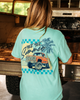 Person wearing a light blue t-shirt with a graphic design of a truck and palm trees, standing in front of a rustic vehicle.
