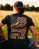Man wearing a black t-shirt with an American flag design and 'Land of the Free' text, sitting in a field.