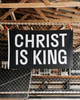 Black banner with 'CHRIST IS KING' text hanging against a wooden and wire fence background