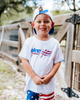 Child wearing a white t-shirt with colorful graphics and American flag pants outdoors.