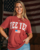 Woman wearing a red 'YEE YEE' t-shirt with an American flag in front of an American flag background.